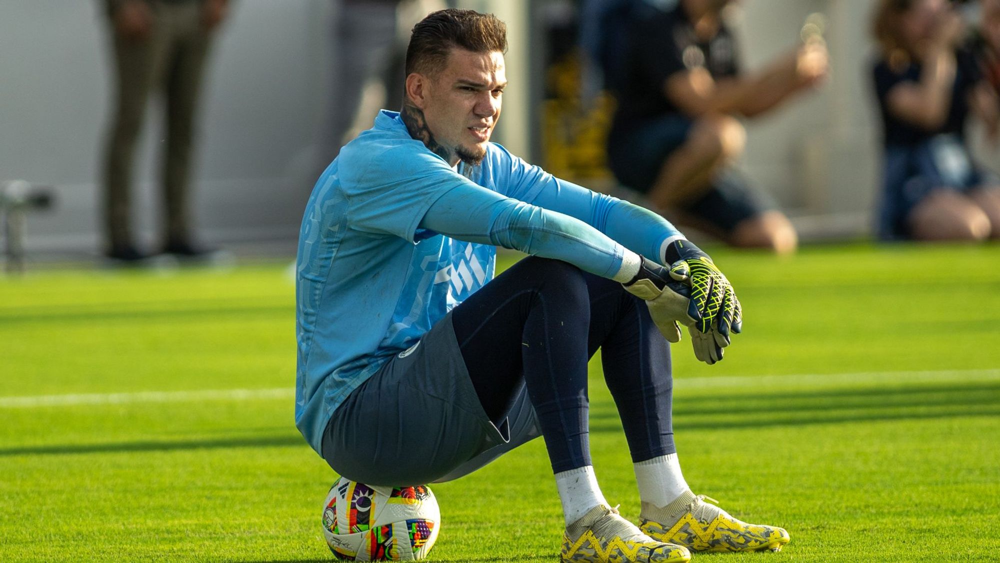 Ederson Sits On A Football Looking On In Training Dressed In A Manchester City Training Kit.